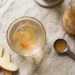 A tablespoon of apple cider vinegar being added to a glass of water, with measuring spoon and ACV bottle nearby